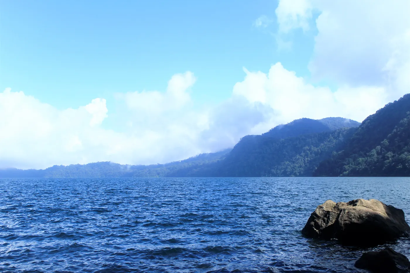 Permukaan Danau Gunung Tujuh yang beriak dengan punggungan bukit berhutan dan awan rendah di kejauhan