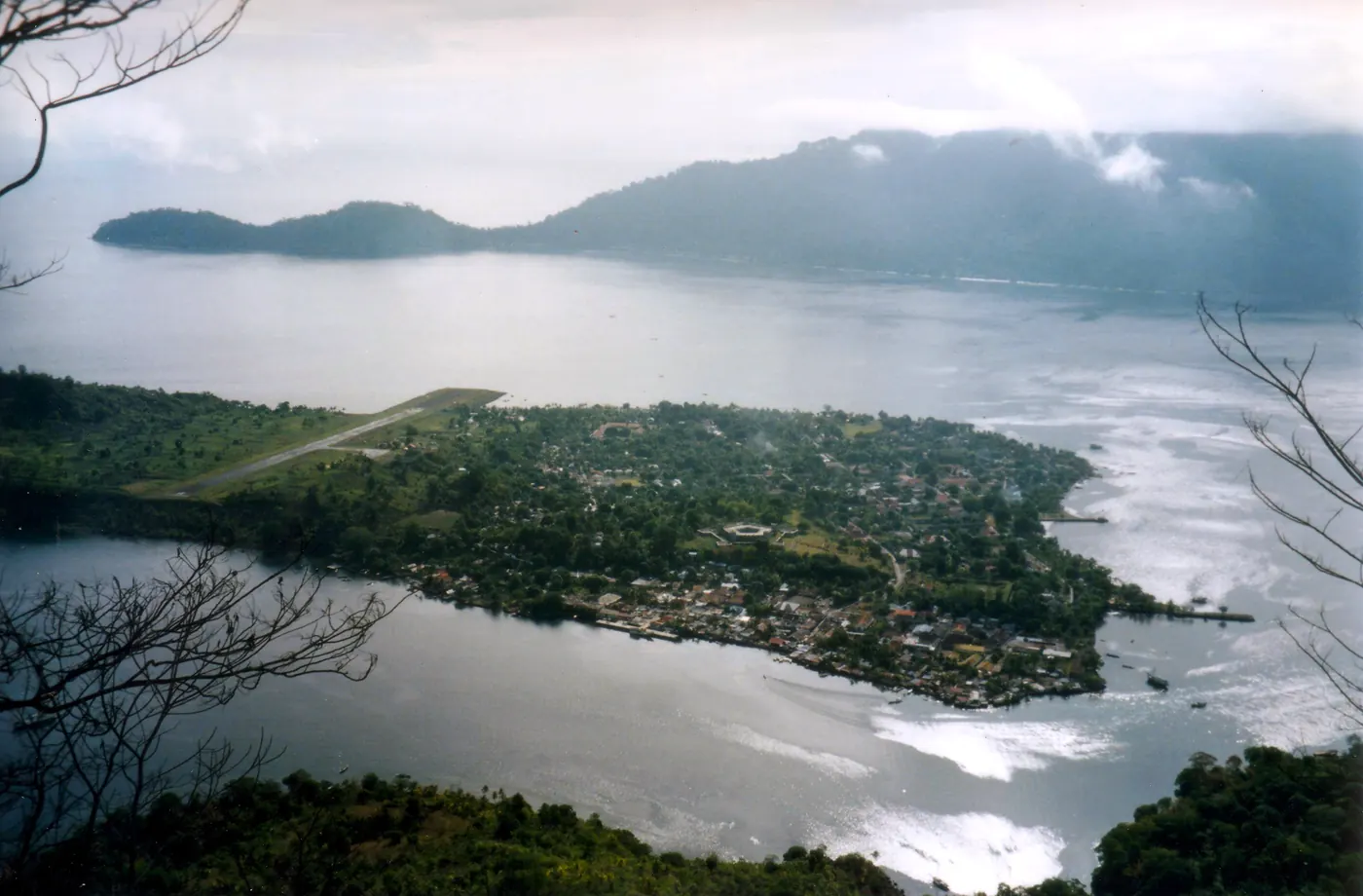 Kota Banda Neira dilihat dari ketinggian Gunung Api, dengan landasan pacu memanjang di tepi kota, selat sempit, dan punggungan Pulau Banda Besar di seberangnya