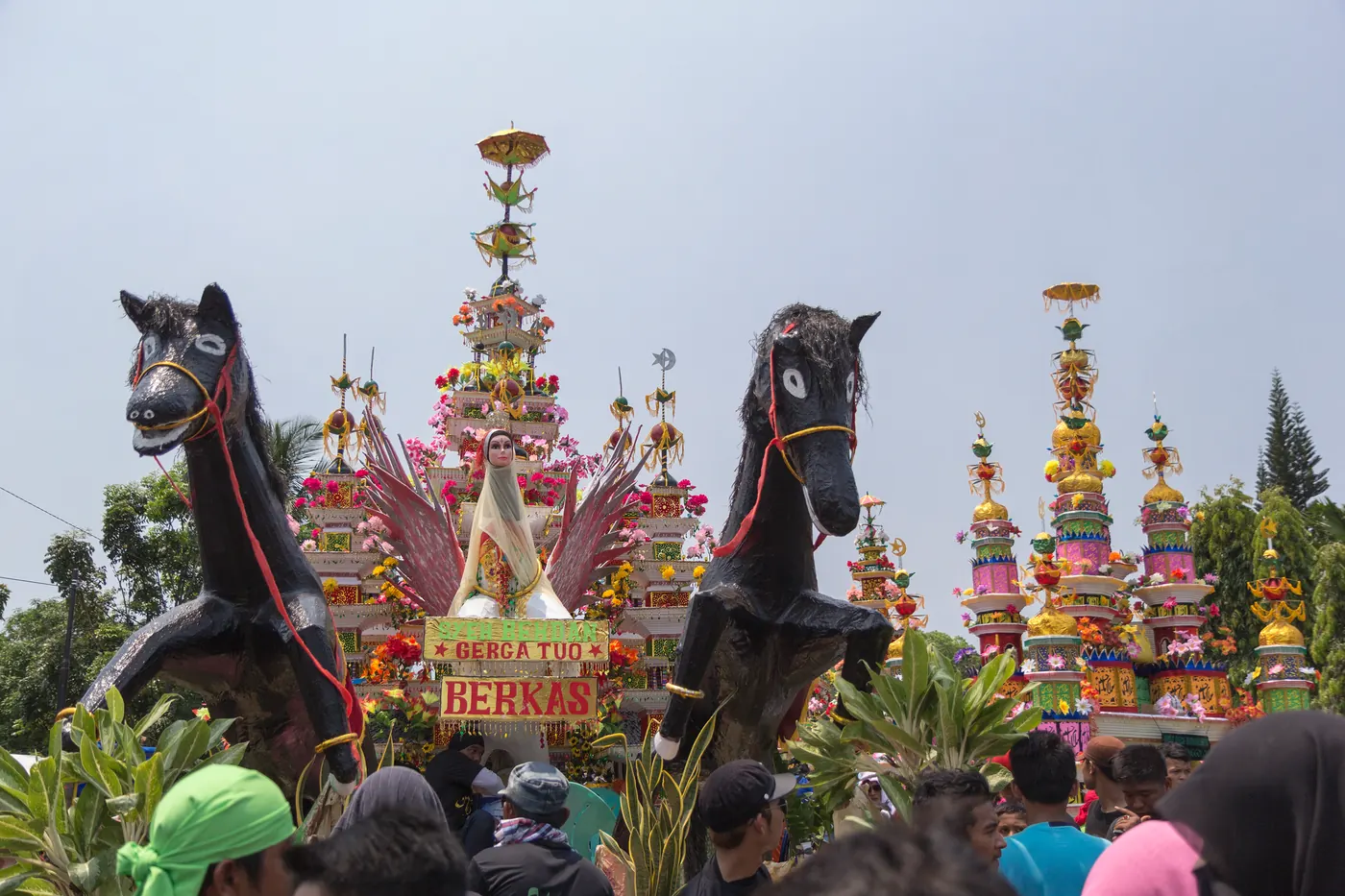 Dua patung kuda bersayap mengapit bangunan Tabot bertingkat berhias bunga, dengan menara-menara Tabot berwarna terang di belakangnya dan kerumunan warga di depan