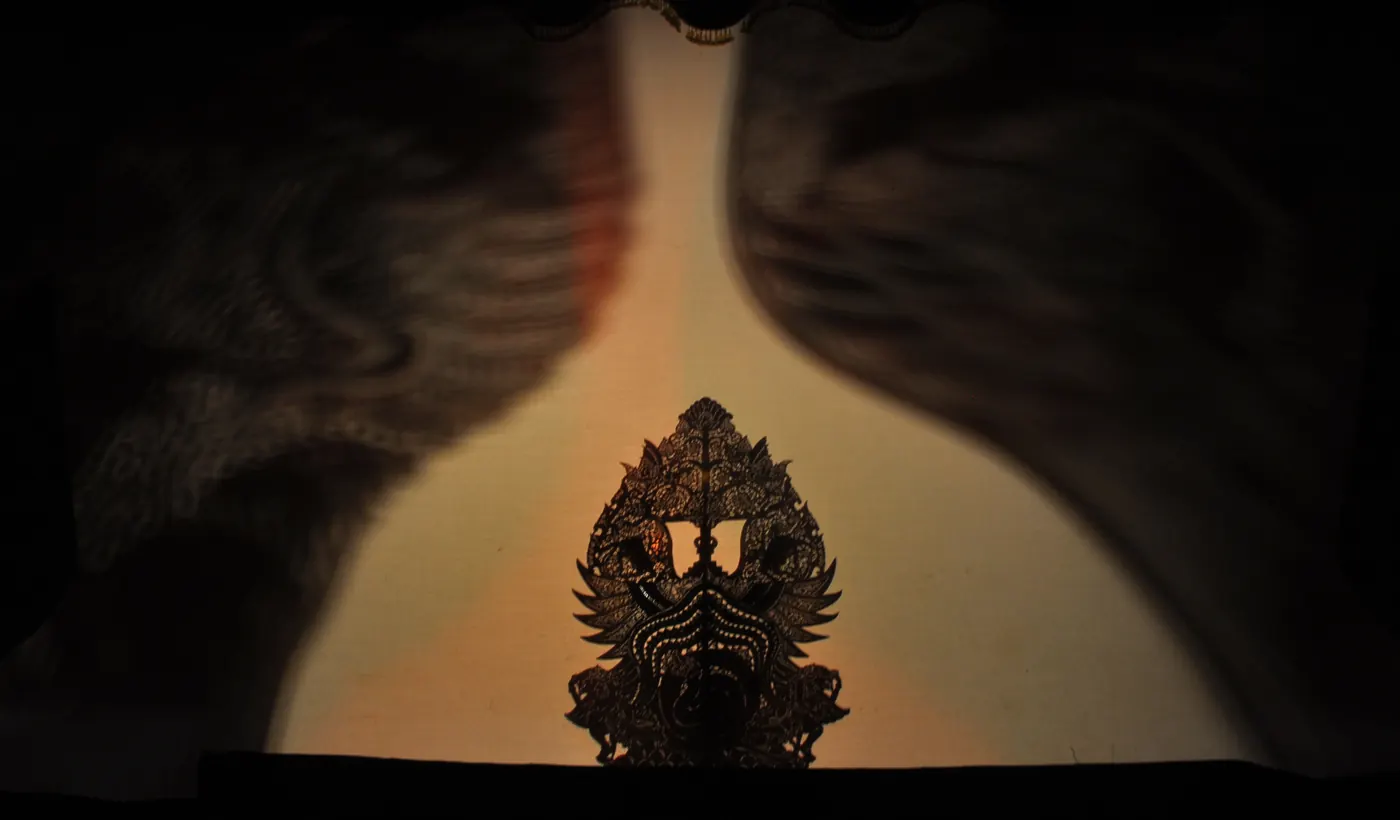 Siluet gunungan wayang tertancap di tengah kelir yang disorot dari belakang, diapit dua bayangan besar yang kabur