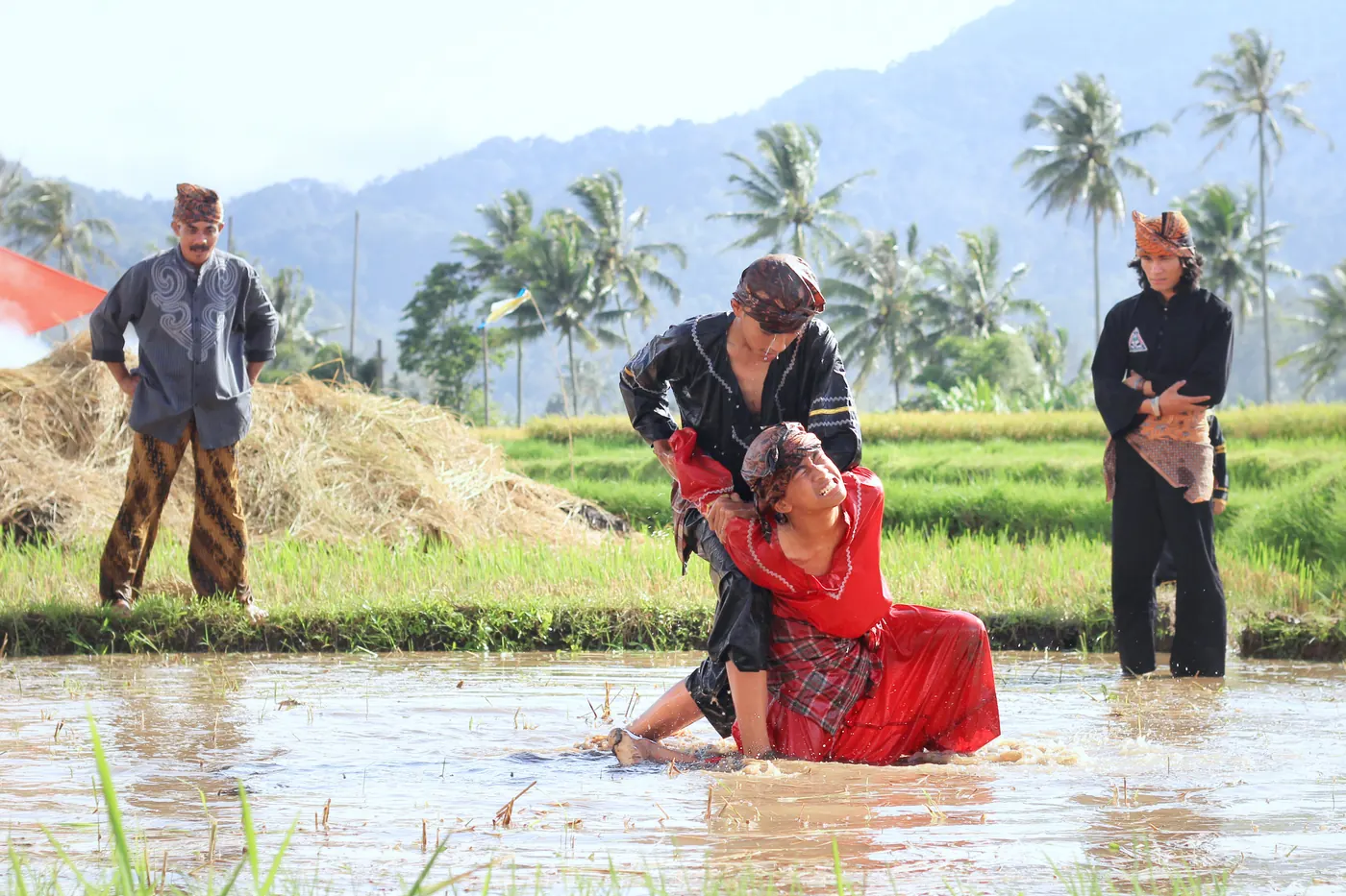 Dua pesilat berbaju hitam dan merah bergerak di sawah berlumpur berair dengan pegunungan di kejauhan dan dua orang lain berdiri mengamati