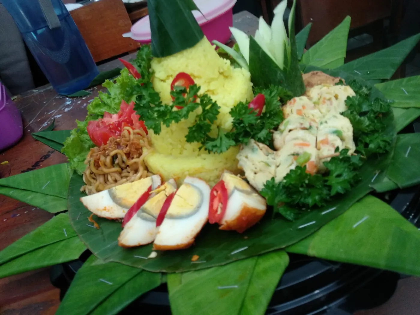 Tumpeng nasi kuning berbentuk kerucut dikelilingi aneka lauk pauk di atas tampah beralas daun pisang