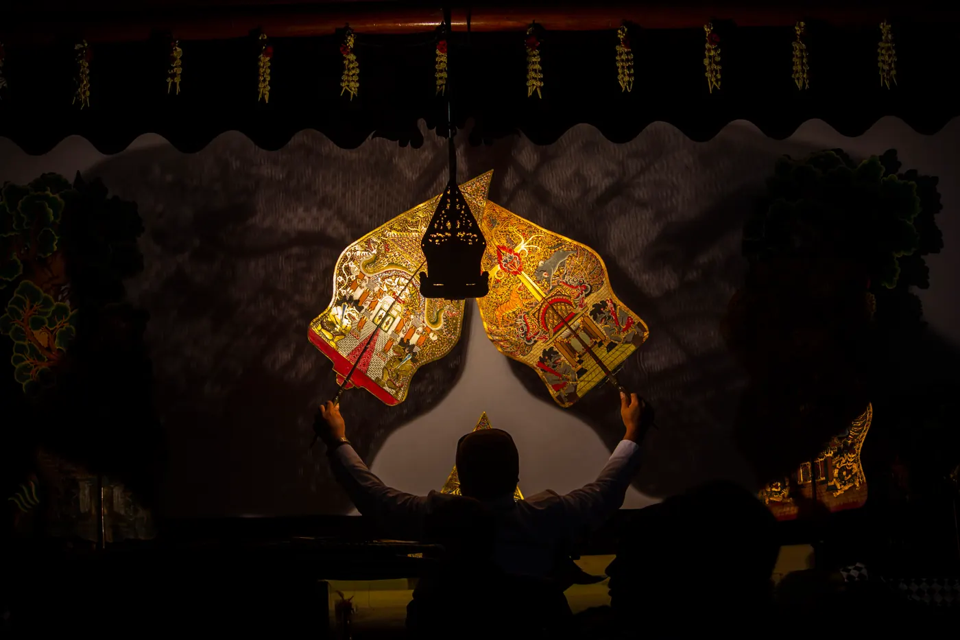 Dalang duduk di depan kelir dengan deretan wayang kulit tertancap di batang pisang dan perangkat gamelan di sekitarnya