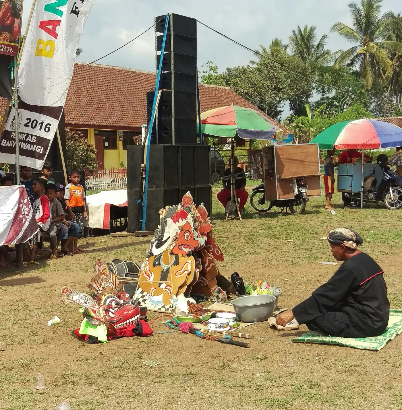 Seorang pawang berbaju hitam duduk bersila di atas tikar menghadap deretan topeng barongan dan wadah-wadah sesaji di sebuah lapangan desa