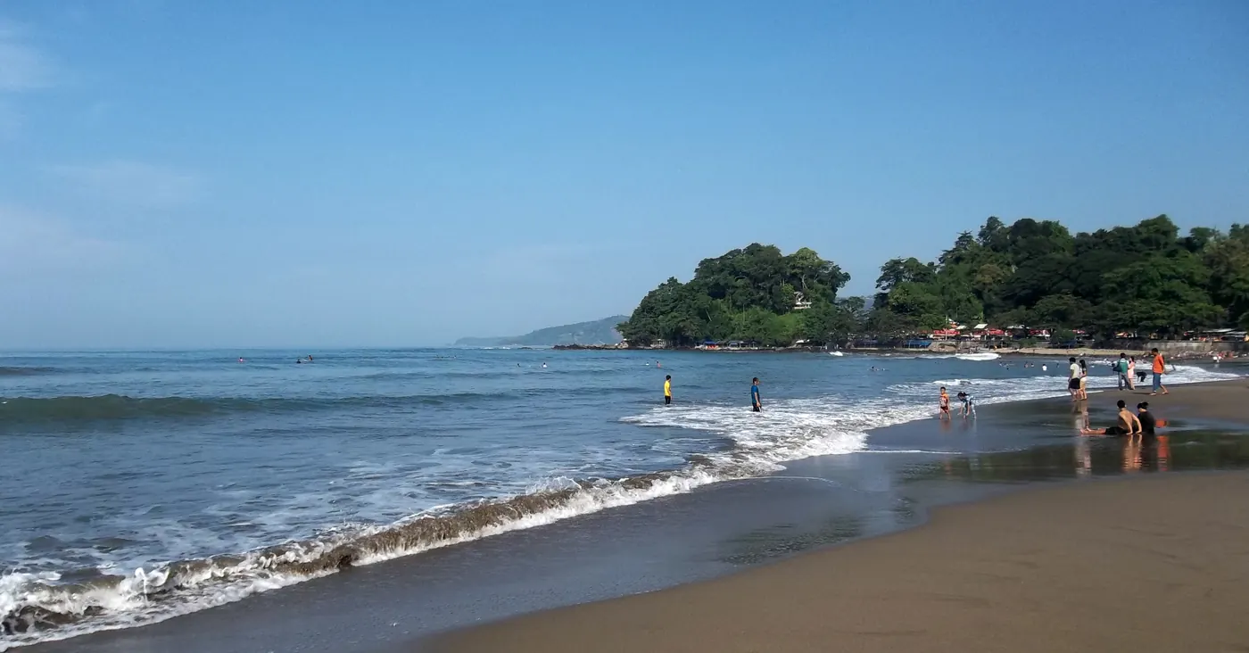 Pengunjung berdiri di air dangkal Pantai Karang Hawu, dengan ombak pecah di garis pantai dan tanjung berhutan di kejauhan