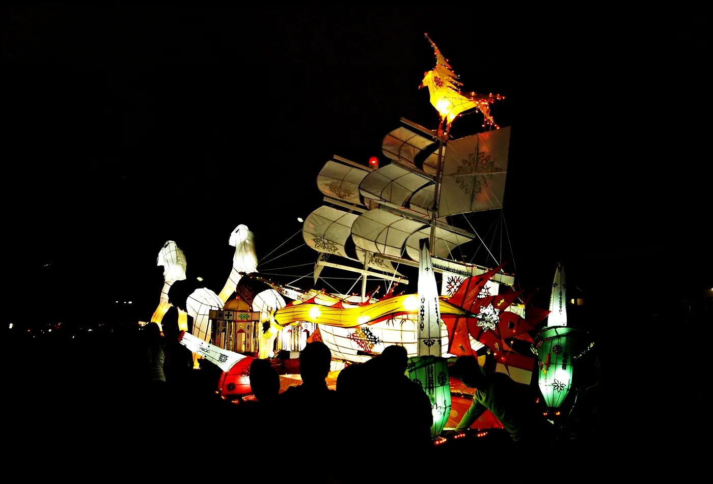 Lampion telong-telong raksasa berbentuk kapal layar dengan burung menyala di puncak tiang, dikelilingi lampion berbentuk ikan dan siluet penonton pada malam hari