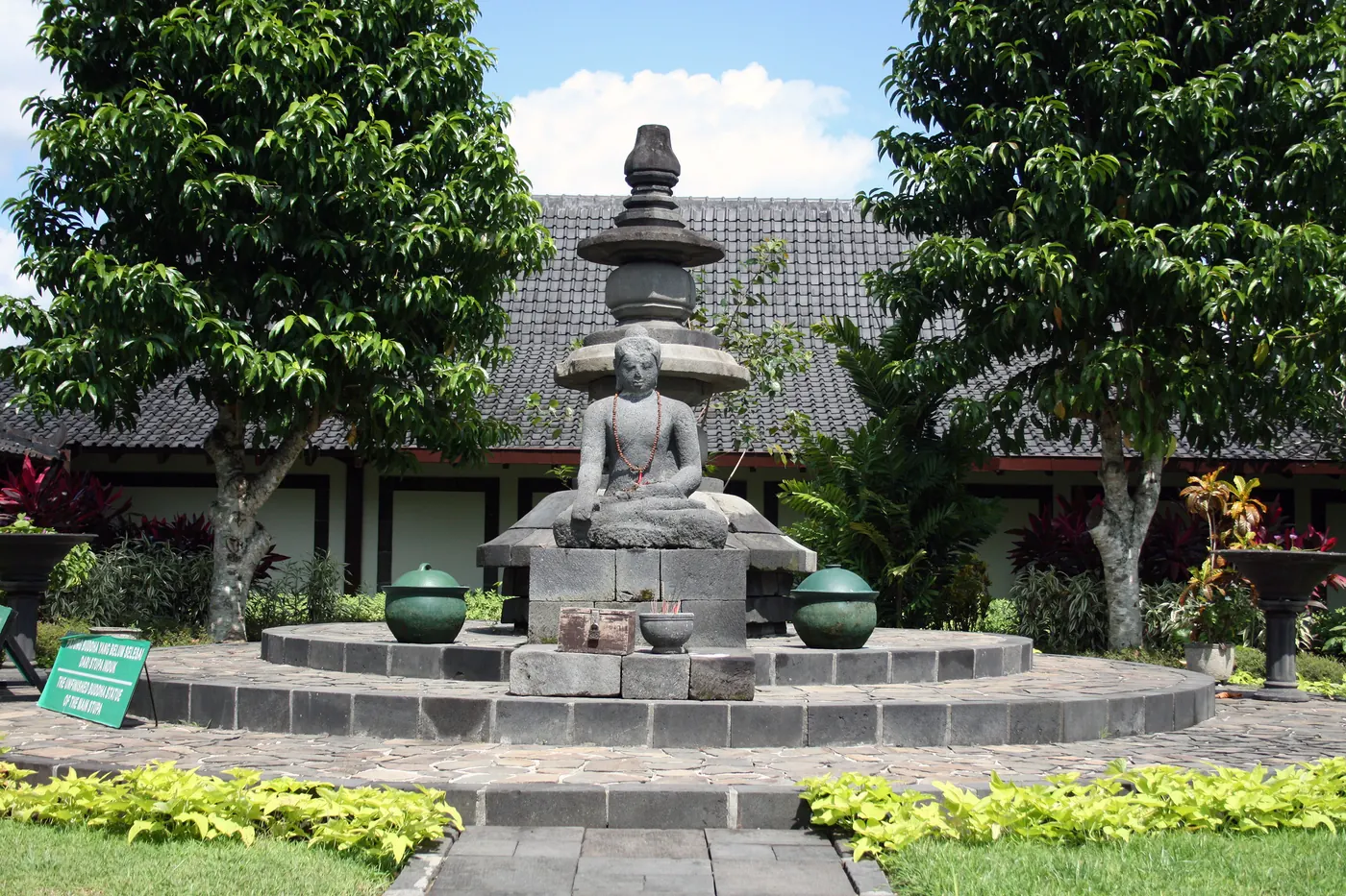 Arca Buddha yang belum selesai dipahat, duduk di alas batu bundar di halaman museum, dengan puncak chatra stupa induk terpasang di atasnya