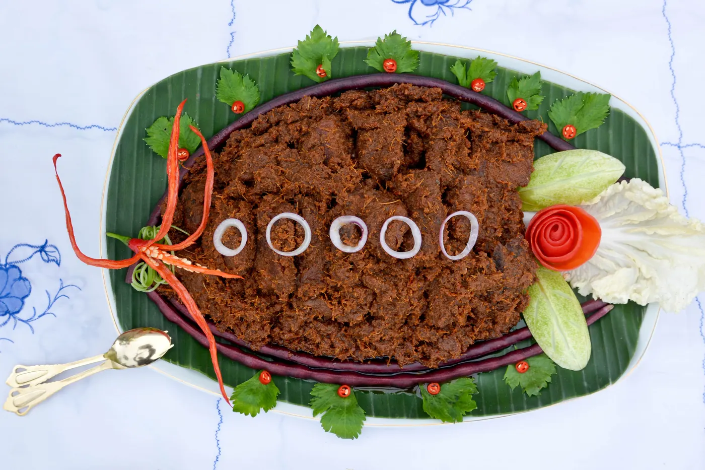 Rendang berwarna cokelat gelap disajikan di atas daun pisang, dihias irisan bawang, cabai merah, dan daun seledri