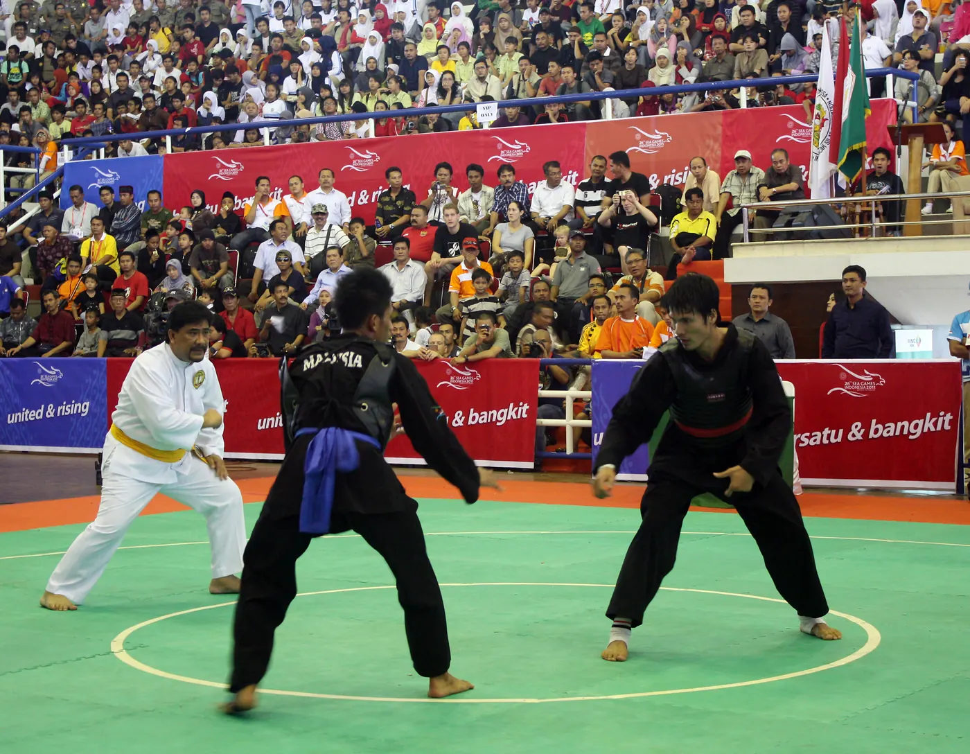 Dua pesilat bertanding di atas gelanggang bertanda lingkaran, dengan wasit dan penonton di sekelilingnya