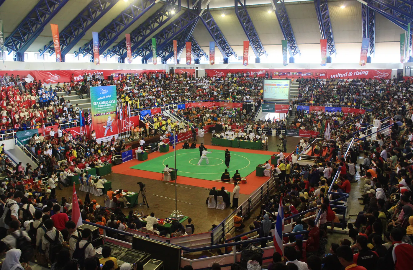 Gelanggang pencak silat bermatras hijau bertepi merah di tengah gedung yang penuh penonton, dengan spanduk SEA Games dan bendera negara peserta
