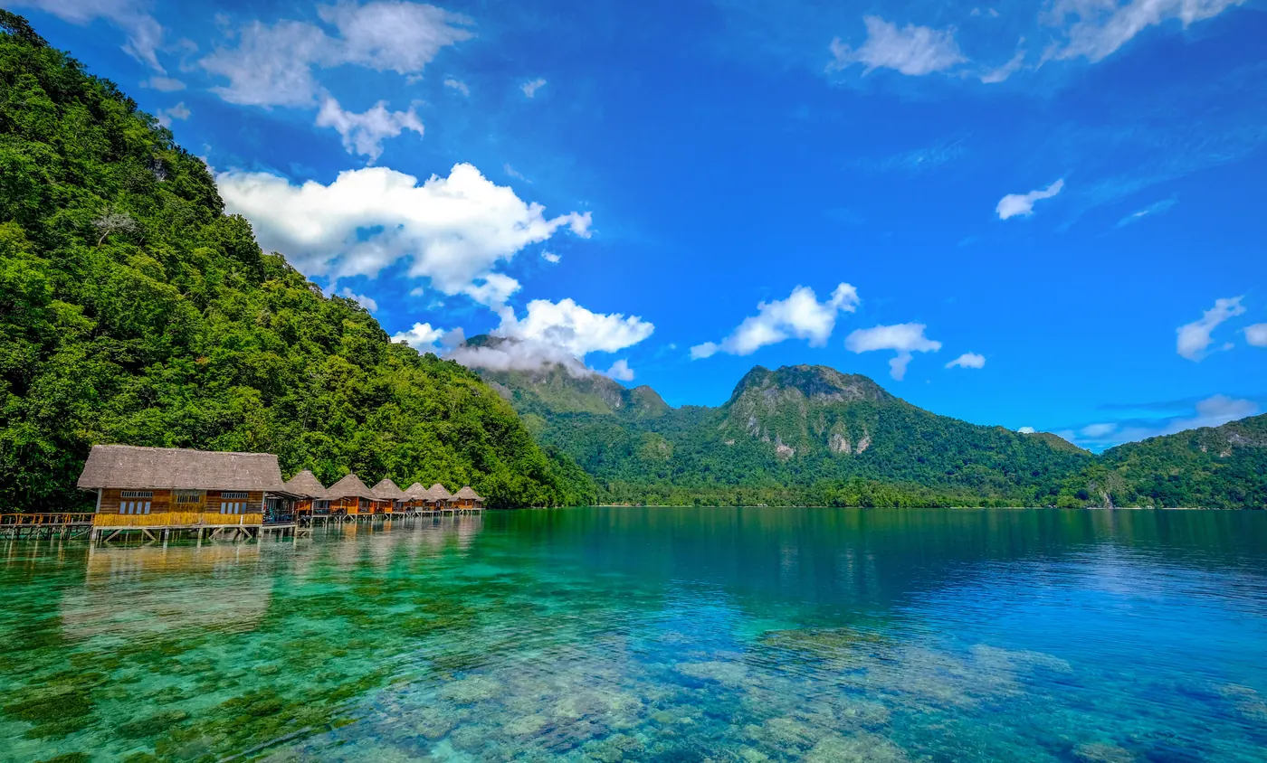 Perairan Maluku dengan air jernih kehijauan, garis pantai berpasir putih, dan perbukitan hijau di kejauhan