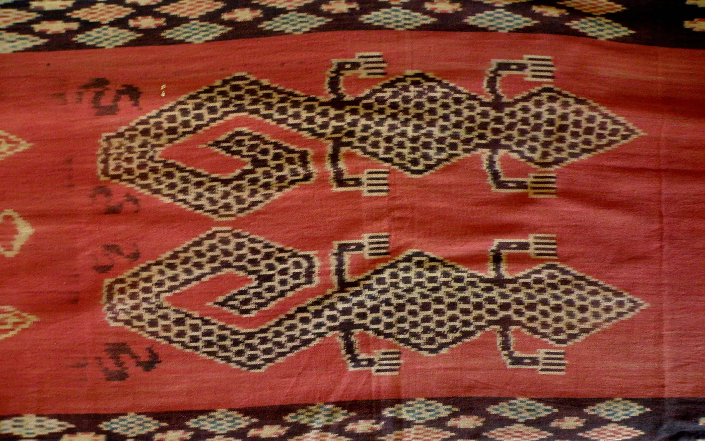 Detail kain hinggi Sumba, memperlihatkan motif figuratif berwarna merah bata dan krem di atas dasar biru kehitaman